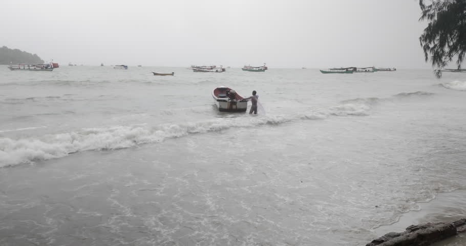 Man struggling with a small motorboat in rough surf during a stormy day,   with several fishing boats in the background.