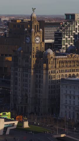Aerial drone sweep shows Royal Liver Building at Pier Head in Liverpool, with clock faces, Liver Bird statue, domes, pilasters, plaza, promenade, and riverside towers.