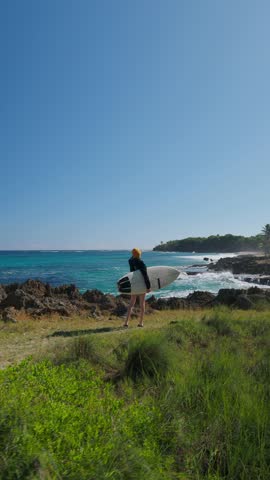 Woman with  surfboard on  tropical surfing spot next to ocean, looking at ocean waves. 