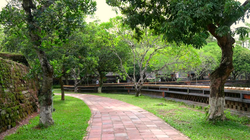 A peaceful brick pathway winds through lush gardens and ancient trees with mossy walls at Tu Duc Tomb, Hue, Vietnam.	