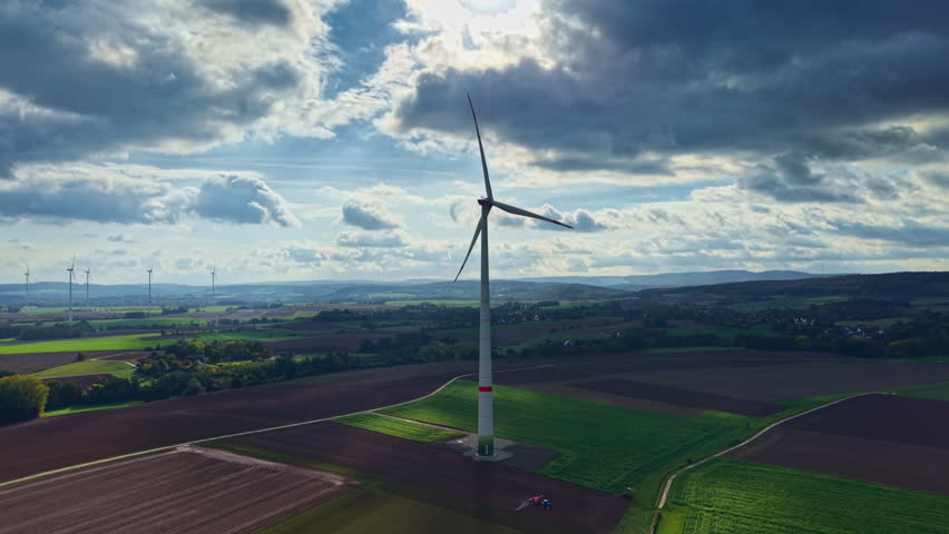 A tall wind turbine stands proudly on rolling farmland as clouds drift overhead The green fields contrast with the brown earth creating a peaceful scene during late afternoon light.