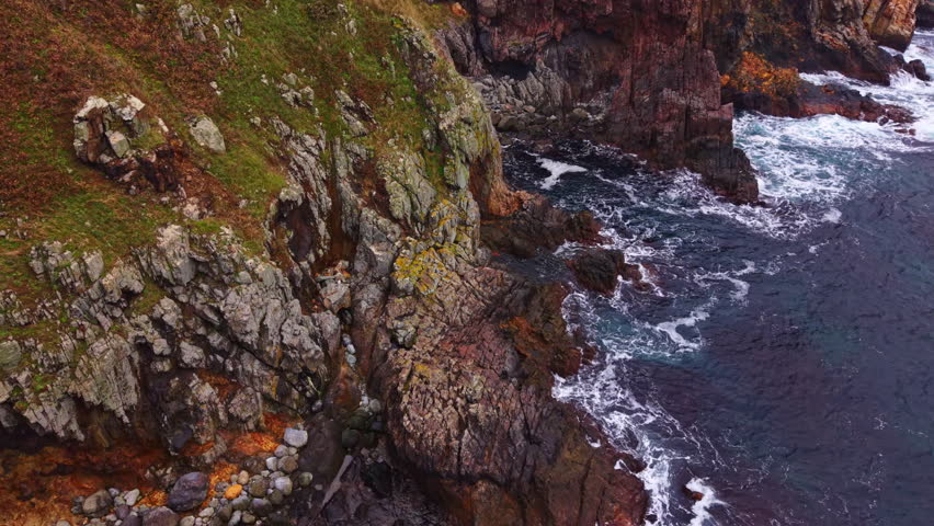 Rugged cliffs tower over the crashing waves as the sun sets on this remote coastal scene. Textured rocks line the shore while gentle waves lap against the stony beach, creating a tranquil atmosphere.