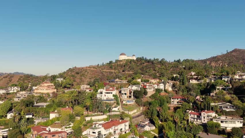 Griffith Observatory overlooking Los Angeles hills and residential neighborhoods under a clear blue sky, aerial drone view.
