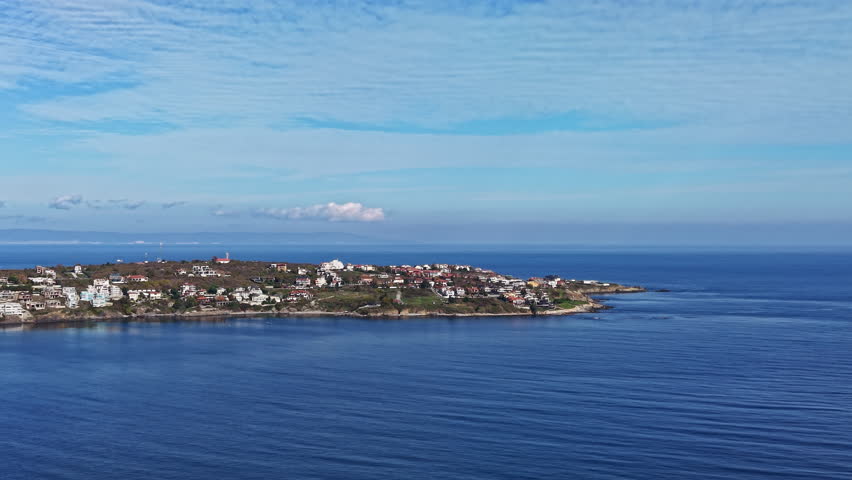 Capturing a serene seaside landscape, the scene features crystal-clear waters gently lapping against the shore while quaint homes dot the coastline under a bright blue sky.
