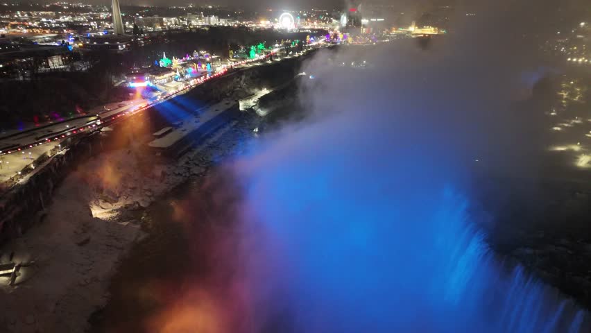 An aerial pulling back to reveal the full edges and powerful water flow of the illuminated American and Horseshoe Falls at night during a winter event