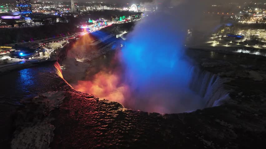 An aerial shot pulling back to reveal the dark, cold water of the Niagara River and the full scope of the illuminated American and Horseshoe Falls at night