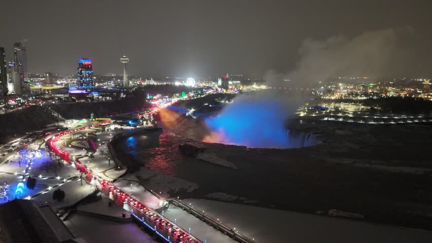 An expansive aerial pull-out shot revealing the full extent of the illuminated American and Horseshoe Falls with vibrant cityscape lights and snow at night
