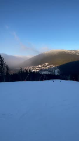 Vertical screen wide snowy ski slope overlooking mountain town and dark pine valley under clear blue sky creating calm winter travel mood with soft evening light