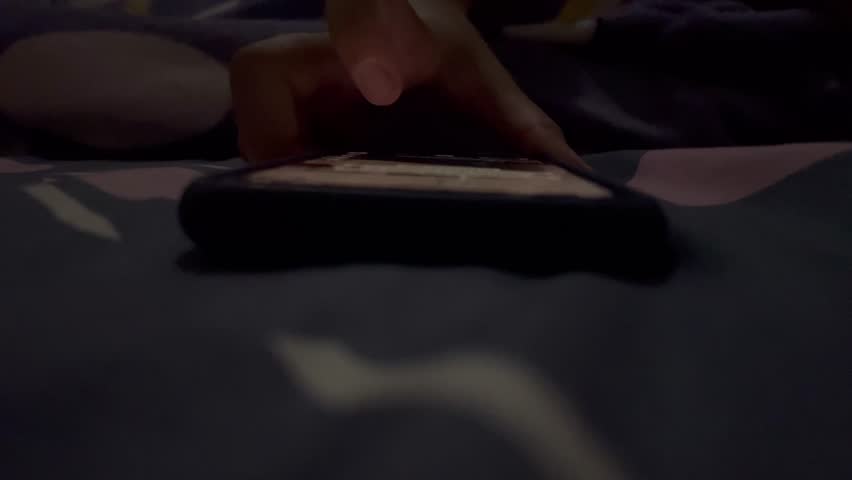 A close-up view of someone playing with their smartphone in their bedroom while they are about to sleep with the lights dimmed.