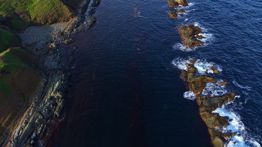 Gentle waves kiss the rocky shoreline as cliffs rise majestically. The sun sets, casting warm hues over the landscape, creating a tranquil coastal scene filled with natural beauty.
