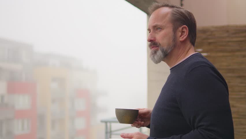 A handsome middle-aged man drinks a cup of coffee and looks out a balcony of an apartment