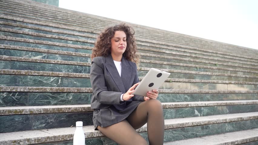 A modern professional woman sits on stone steps using a tablet, showcasing the balance between work and life while enjoying an outdoor setting that sparks her productivity and creativity