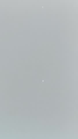 A serene sky view capturing two distant aircraft at high altitude, appearing as tiny white silhouettes against a soft grey sky. The minimal composition highlights the vastness of the atmosphere and th