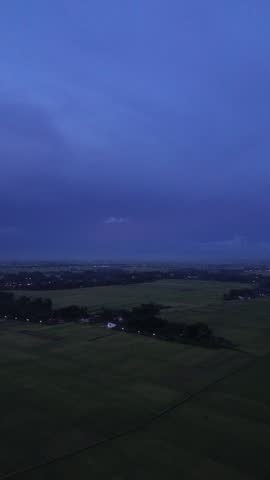 Cinematic aerial drone footage of a stormy blue hour sky dominating the frame, with distant lightning glowing behind thick clouds above a rural farmland landscape. The darkening fields and scattered village lights below contrast with the deep blue atmosphere, creating a dramatic and moody evening scene. This footage represents approaching storm, weather changes, nightfall transition, and emotional natural landscapes. Ideal for cinematic openers, weather-related projects, documentaries, dramatic transitions, environmental storytelling, and atmospheric background visuals. Clean composition with no people, logos, or text, suitable for commercial stock usage.