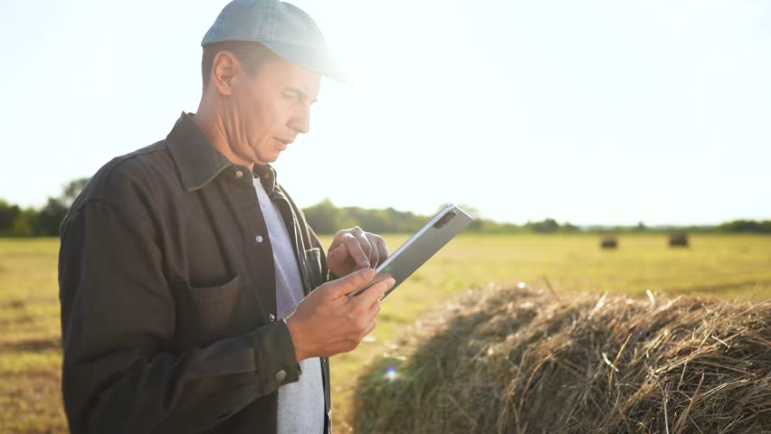 farmer man with a cap uses the tablet beside the hay bale in the field. The man focuses on the tablet and stands near the hay bale under the open sky in the field.