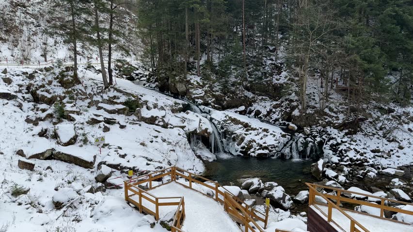 A scenic winter landscape features a mountain waterfall cascading into a clear pool, surrounded by snow-covered rocks, large icicles, tall evergreen trees, and wooden observation decks