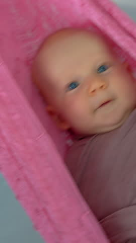 Loving mother gently swings her swaddled two-month-old baby in a hammock to help him fall asleep, creating a peaceful and nurturing family bedtime moment.