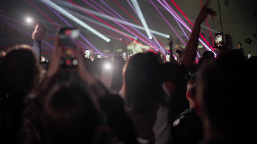 Concert crowd raising smartphones and jumping while filming a live performance under laser stage lights