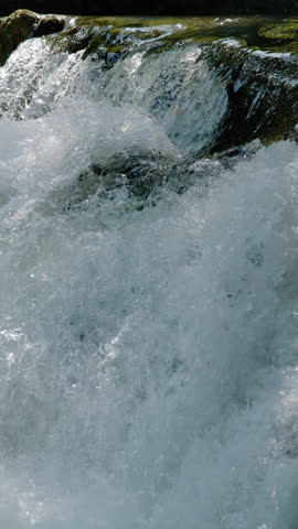 VERTICAL, CLOSE UP, SLOW MOTION: White foam and turbulent movement of crystal clear water cascading down a rocky riverbed. Sunlight highlights the dynamic natural patterns of a free flowing river.