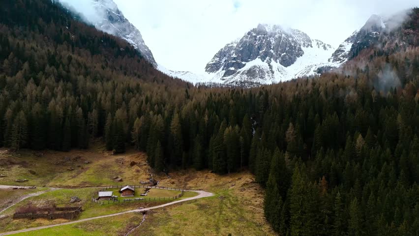 Cinematic drone footage of the Italian Alps. Snow-capped peaks rise over a dense conifer forest and a small group of wooden alpine cabins with a fenced meadow in a tranquil valley. Low clouds and light mist drift across the slopes. No people or logos visible. Property is identifiable, so this clip should be licensed as editorial.