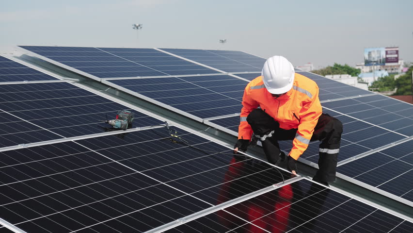 Technician installing solar panels on factory roof for green energy. A skilled technician in safety gear is working on a solar panel installation on rooftop.