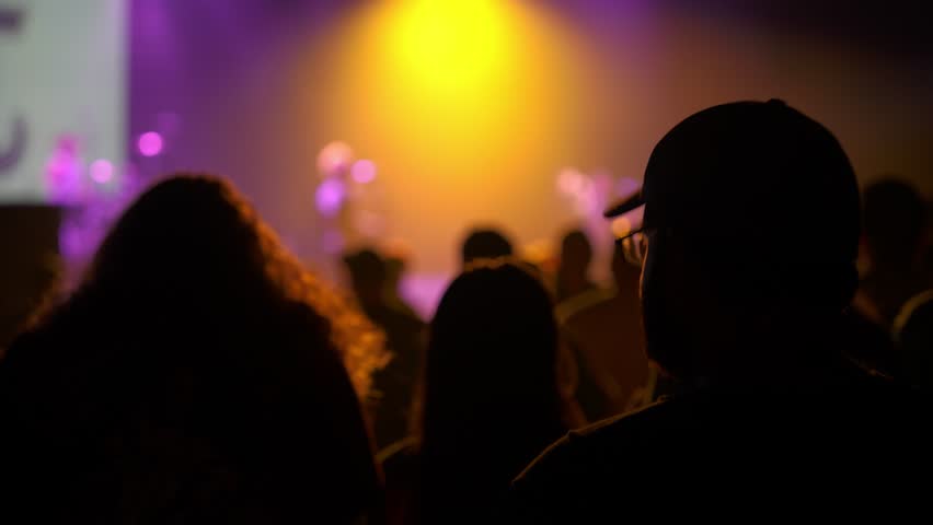 Dark silhouettes of concertgoers watch a performance with a bright yellow spotlight on stage. Purple lighting, haze, and bokeh create a dramatic live music mood.