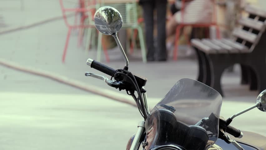 Motorcycle front with handlebar, mirrors, and windshield shown in foreground. Cafe chairs, bench, and pedestrians appear blurred in background.