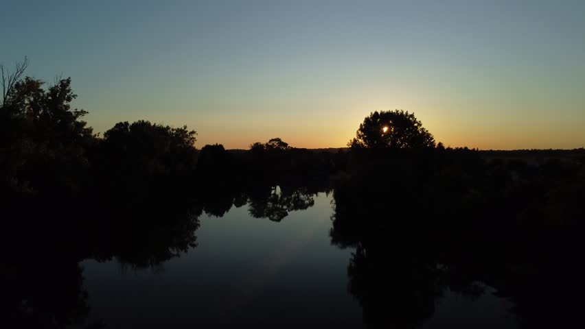 The sun sets behind trees as the sky changes colors over a calm river. The area is remote, highlighting nature and the beauty of the evening light.