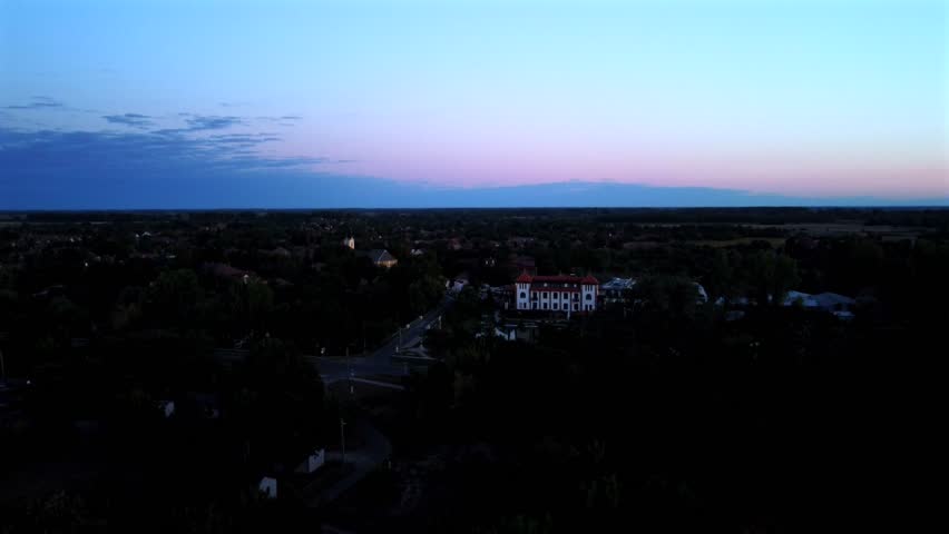 The camera captures a view of a small town at dusk. Buildings and trees are visible as the sky changes color. The layout of roads and houses shows the town's layout and surroundings.