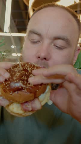 Vertical Screen: A man bites into a burger with excitement at a modern restaurant. The atmosphere is lively, and the flavors promise a satisfying meal.