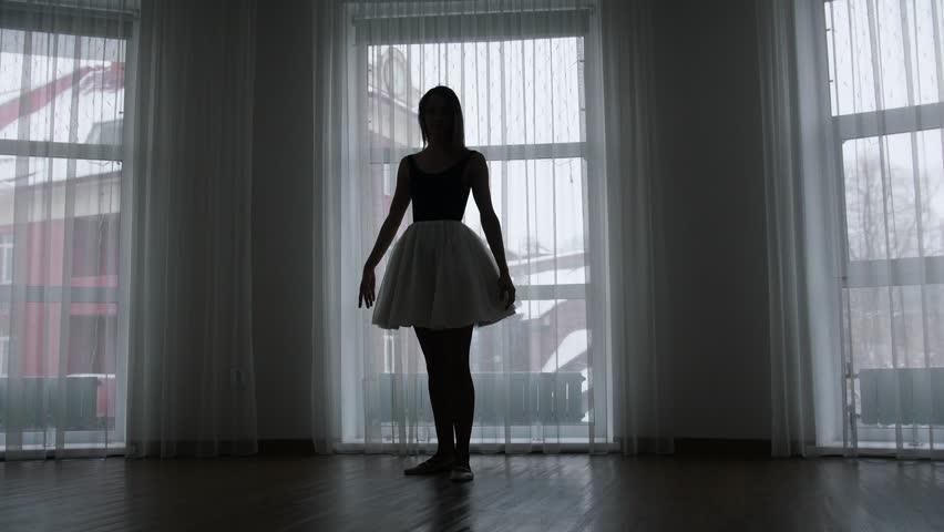 A studio in twilight. A silhouette of young woman ballerina training her spinning in front of the window