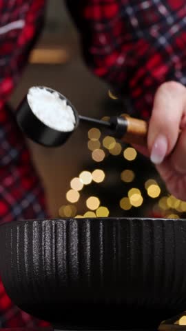 Woman adding starch into a black bowl with milk, showcasing the preparation of a hot chocolate drink with festive background lights