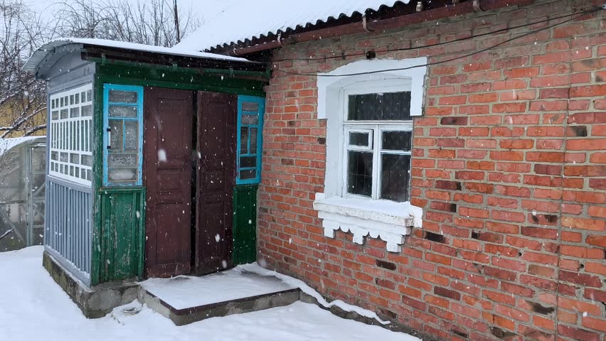 A serene winter scene features a weathered brick house with a white-framed window, adjacent to a small green outbuilding with blue-trimmed doors. Snow blankets the ground, and gentle snowfall creates 