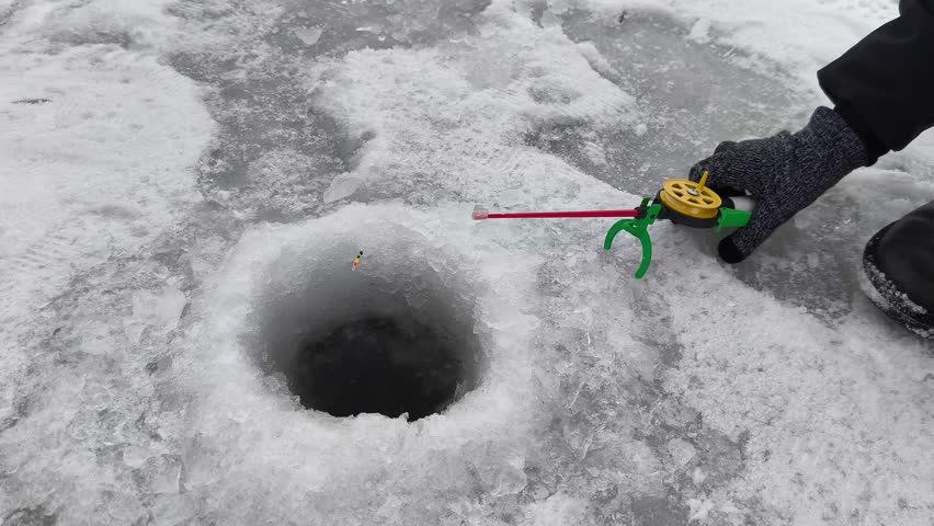 Close-up view captures an ice fishing scene where a person uses a green and yellow reel to catch a fish through a hole in the frozen lake. Snowy surroundings highlight winter fishing activity, showcas