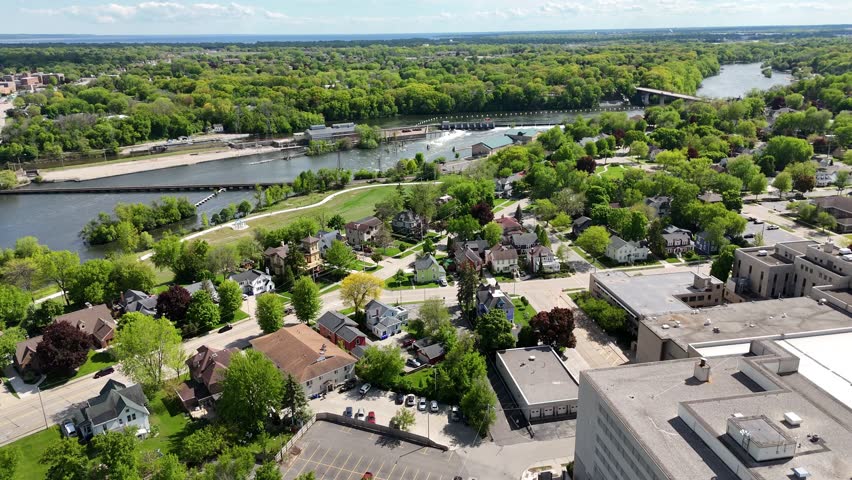 High push Drone shot approaching Fox River in Appleton Wisconsin USA.  Dam and Bridge in the distance.  Blue Skies, with scattered clouds through the sunshine a warm summer day. Downtown Hugs riverside. Shadows linger over the landscape. 