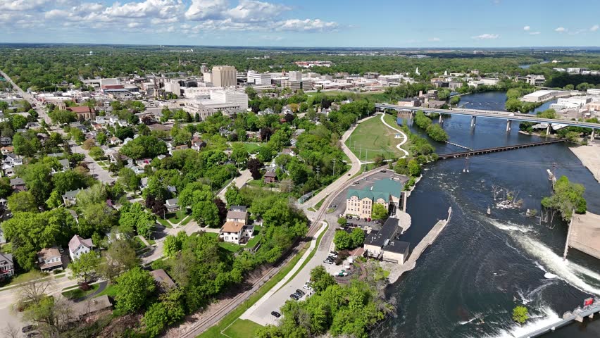 High push Drone shot approaching Bridge over Fox River in Appleton Wisconsin USA.  Dam and Bridge in the distance with Cars driving.  Blue Skies, with scattered clouds through the sunshine a summer day. Downtown Hugs riverside. Cloud shadows linger over the landscape.