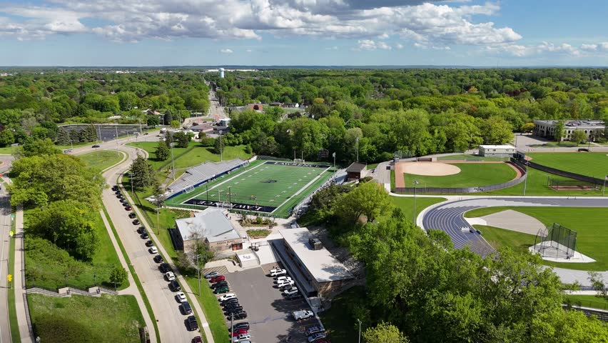 Slow Drone push shot over Athletic complex and College Football Field.  Spectators and players on the field.  Car filled streets below and blue cloudy Skies above. Appleton Wisconsin USA