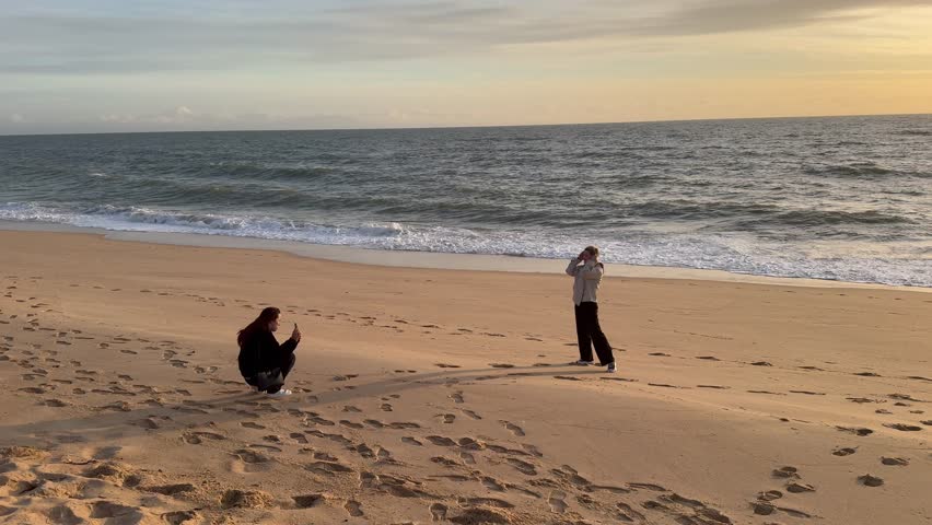 Mobile photo session of two travelers on seacoast in sunset, on beach. Young adult woman posing on shore and girl photographing her, slow motion, backlit shot, local tourism