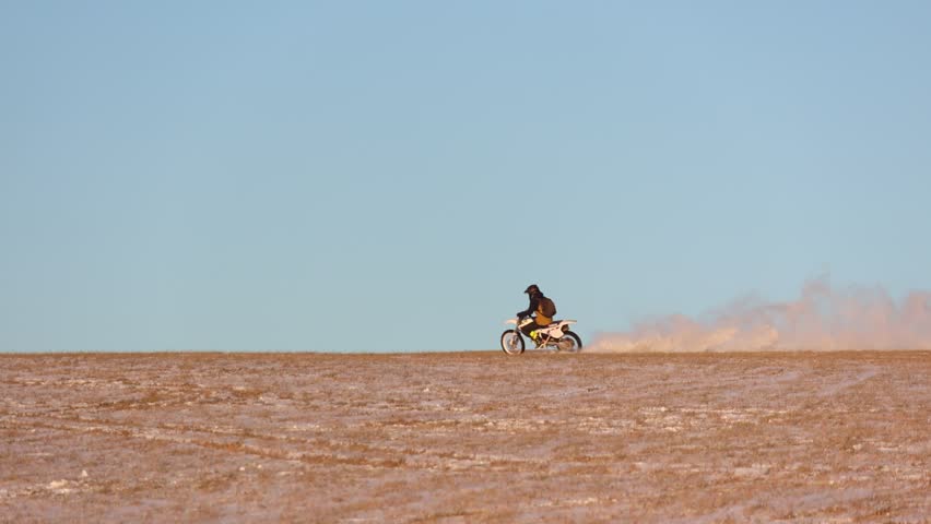Dirt biker speeds across a sprawling, snow-dusted field on a bright, sunny day, kicking up a cloud of dust, showcasing the excitement of off-road riding and the expansive beauty of nature.