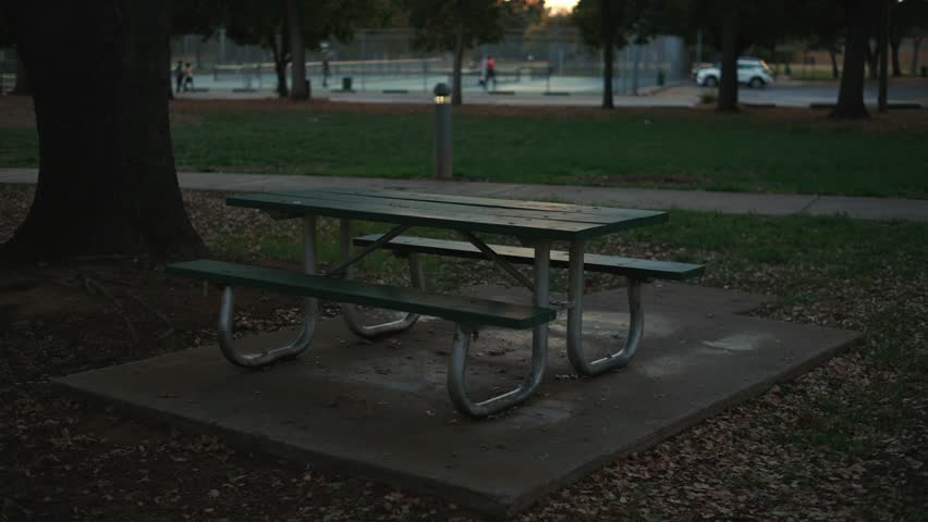 Empty picnic table near tennis courts in a city park at sunset, green wooden surfaces and metal supports on a concrete pad among fallen leaves
