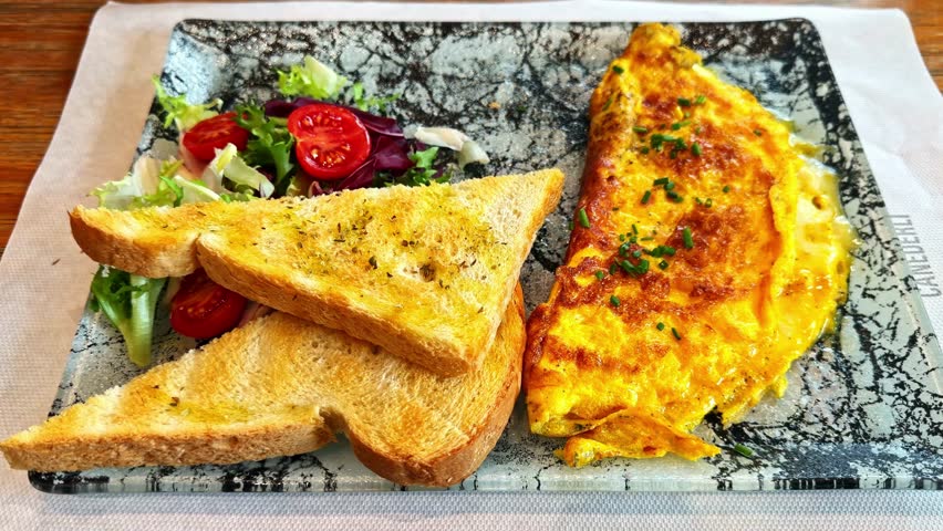 Golden omelette served with toasted bread and a fresh salad mix, captured on a rustic plate in a cozy breakfast setting.