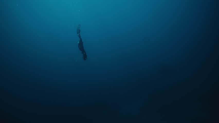 Solo free diver descends into deep ocean depths as dark silhouette in slow motion