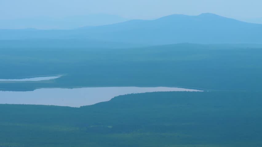 A serene, atmospheric shot of a vast lake surrounded by dense forests and rolling mountains in the Southern Urals region of Russia. The landscape is bathed in a cool, monochromatic blue and teal color