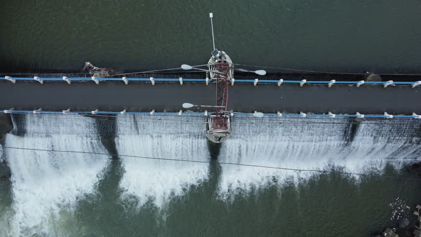 Drone view of Katulampa Water Gate in Bogor, West Java, Indonesia, showing water cascading over a spillway as motorcycles cross the bridge above the dam. This reservoir functions as an important water control point managing river flow from the Puncak area to Jakarta. The scene highlights the interaction between public infrastructure, water management systems, and daily transportation activity.