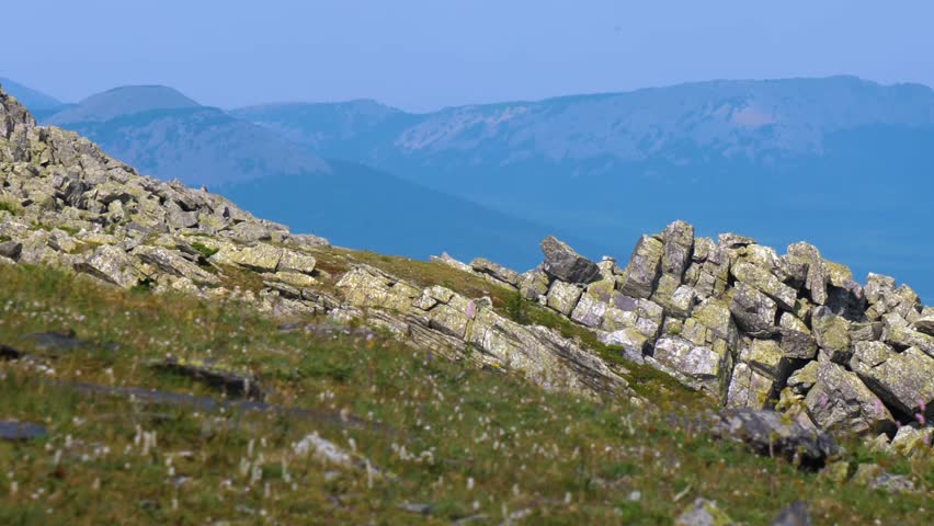 A scenic view of a mountain landscape in the Southern Urals, Russia. The foreground features a steep, grassy slope dotted with large, weathered rock formations and boulders. In the background, rolling