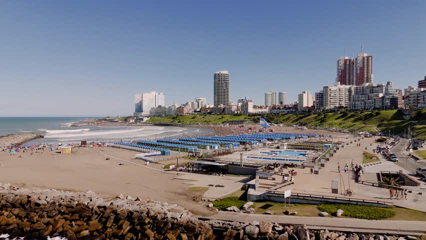 Sunny day view of the crowded Varese beach in Mar del Plata, Argentina, featuring beach umbrellas, tents, and a flying Argentinian flag. The camera is static