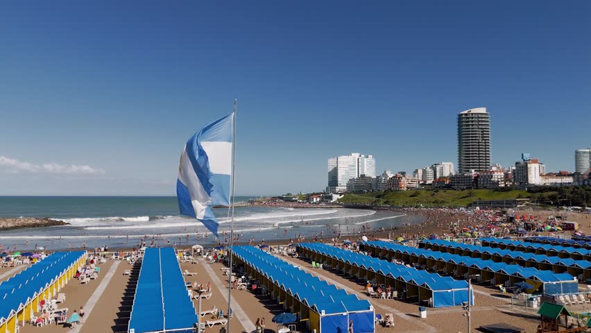 Aerial view of the Argentinian flag in the foreground and crowded Varese beach in Mar del Plata, Argentina. The camera performs an orbit movement
