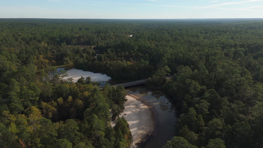 Smooth aerial pass above bridge as river meanders creating sandy beaches by dense green forest in Blackwater River State Park