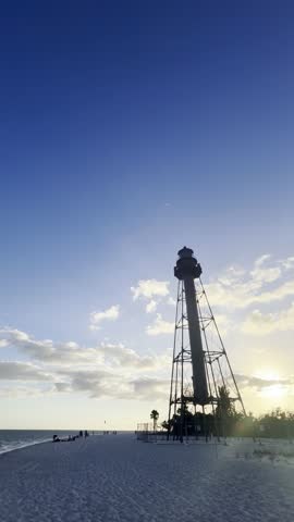 Sanibel Island Lighthouse silhouetted against a vibrant twilight sky and scattered clouds, overlooking the sandy beach with people enjoying a peaceful summer vacation in Florida, United States