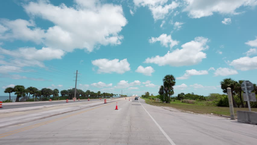 POV driving footage on a coastal highway under a clear blue sky. Road and construction markings, green trees.